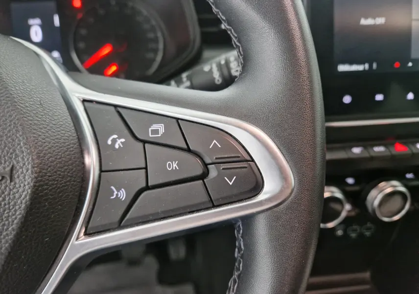 Gros plan sur les commandes du volant côté droit d'une Renault Clio V gris platine, avec tableau de bord et écran tactile en arrière-plan.