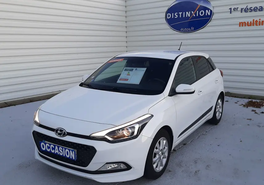 Vue 3/4 avant droite d'une Hyundai i20 blanche de 2016 avec phares allumés, stationnée devant un mur blanc.