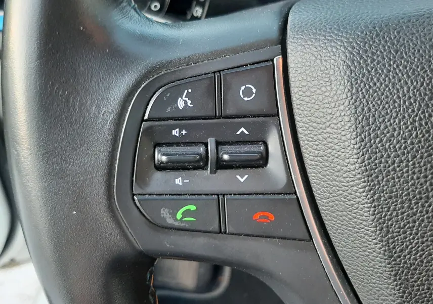 Gros plan sur les commandes multifonctions noires du volant d'une Hyundai i20 blanche, avec boutons d'appel et réglages audio.