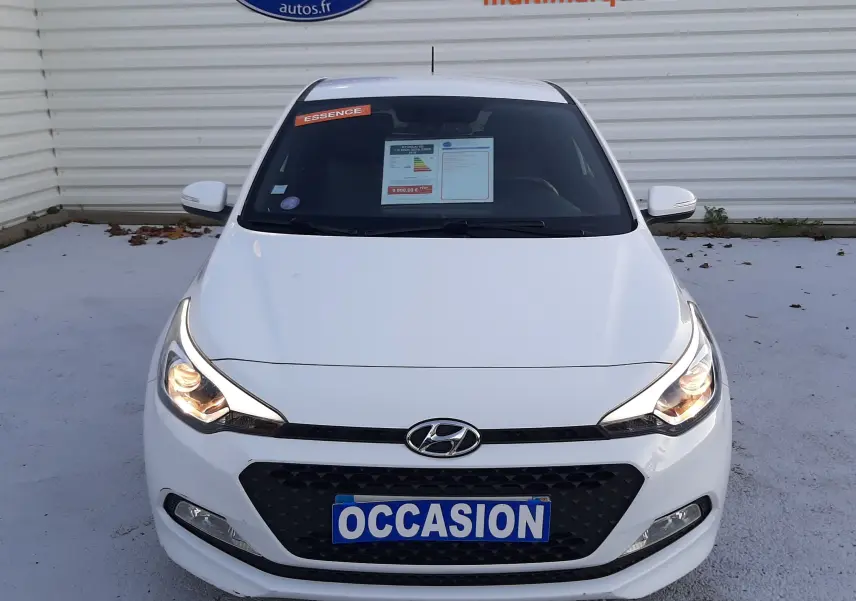 Vue avant d'une Hyundai i20 blanche de 2016 avec phares allumés et plaque "OCCASION" visible sur un parking.