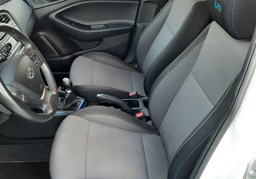 Vue intérieure côté conducteur de la Hyundai i20 blanche, sièges tissu gris avec logo i20 brodé en bleu sur les appuie-têtes.