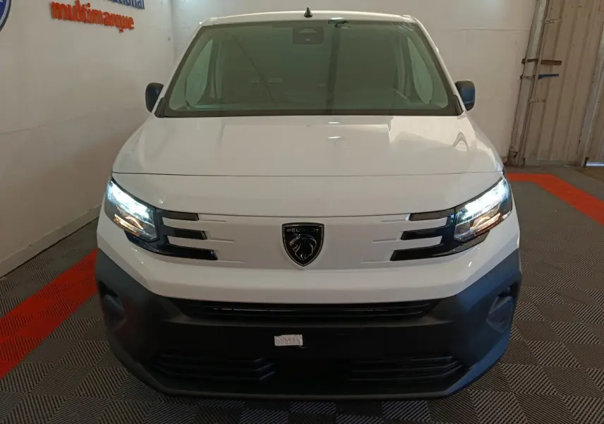 Vue frontale d'un Peugeot Partner blanc 2025 avec phares allumés et logo lion bien visible sur la calandre.