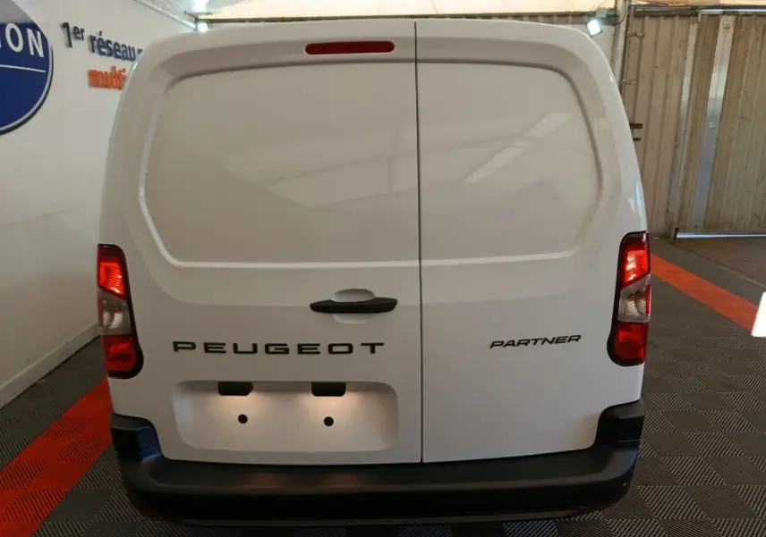 Vue arrière d’un utilitaire blanc Peugeot Partner avec portes battantes et feux arrière allumés dans un garage.