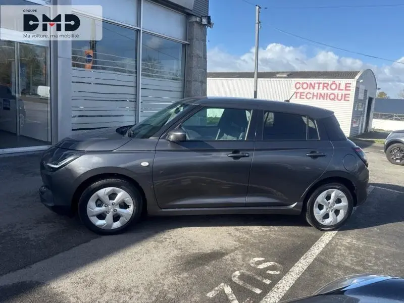 Vue de profil côté gauche d'une Suzuki Swift 1.2 Hybrid 83ch Privilège gris minéral métallisé, garée devant un bâtiment.