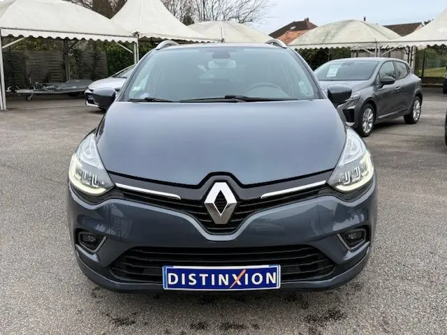 Vue avant d'une Renault Clio IV Estate gris titanium avec phares allumés et plaque d'immatriculation Distinxion visible.