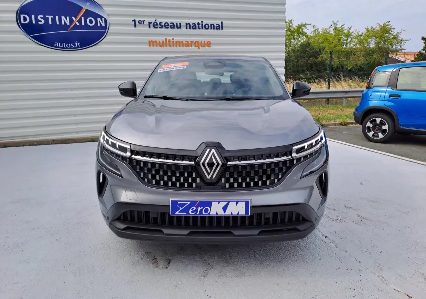 Vue frontale du Renault Austral gris schiste avec calandre à damier et feux LED distinctifs, stationné devant un mur publicitaire.