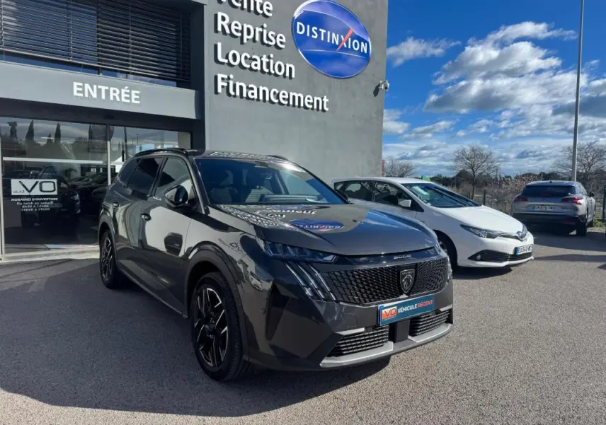 Peugeot 5008 Hybrid gris en 3/4 avant droit, avec jantes alliage noires et calandre distinctive, devant un showroom.