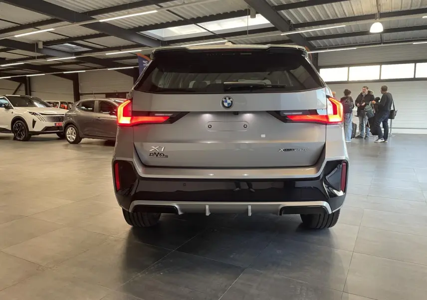 Vue arrière d'un BMW X1 xDrive 20d 2024 en gris Spacesilber Metallic avec feux arrière LED allumés en intérieur showroom.