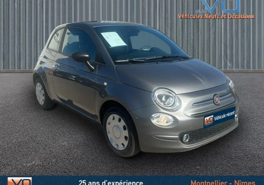 Vue 3/4 avant d'une FIAT 500 gris métallisé 2023 avec phares allumés sur fond urbain neutre.