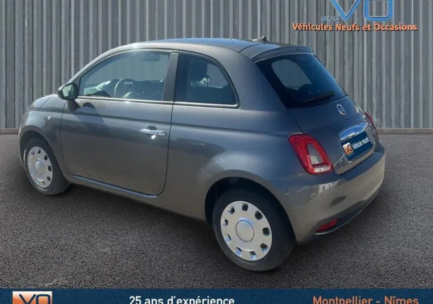 Fiat 500 gris vue 3/4 arrière côté gauche, compacte et moderne sur fond urbain neutre.