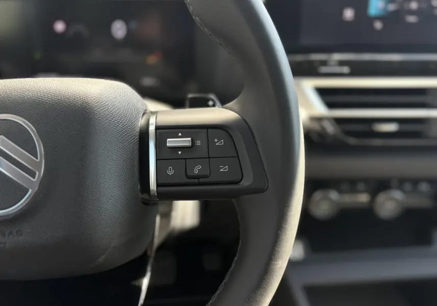 Gros plan sur les commandes au volant gauche du Citroën C4 Hybrid 145 e-DSC6 Max, intérieur noir avec écran tactile flou en arrière-plan.