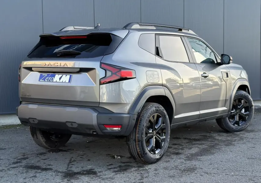 Vue 3/4 arrière droite du Dacia Duster 2026 gris schiste avec jantes alliage noires et barres de toit longitudinales.