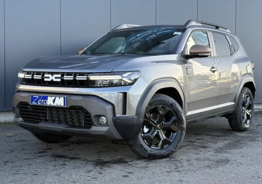 Vue 3/4 avant droite du Dacia Duster 2026 gris schiste avec jantes alliage noires et barres de toit longitudinales.