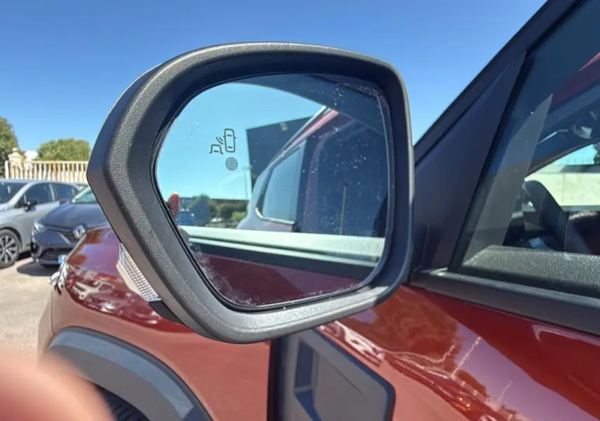 Gros plan sur le rétroviseur côté gauche du Dacia Duster orange 2025, avec capteur d'angle mort visible.