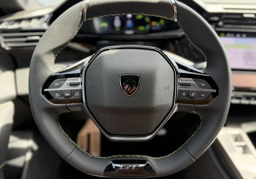 Gros plan sur le volant compact GT en cuir noir avec surpiqûres vert Adamite et logo Peugeot central.