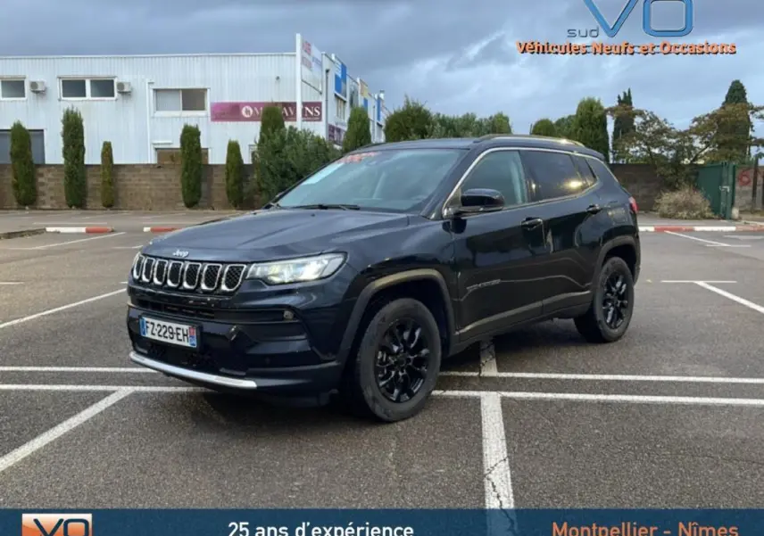 Jeep Compass noir en 3/4 avant droit sur parking, avec jantes alliage noires et barres de toit visibles.