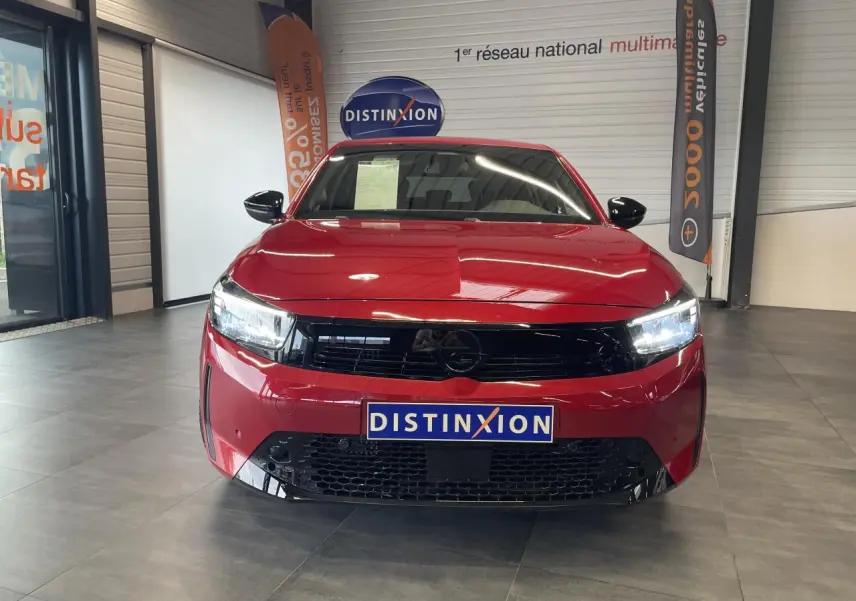Vue avant d'une Opel Corsa rouge 2026 avec calandre noire brillante et phares LED allumés en intérieur showroom.