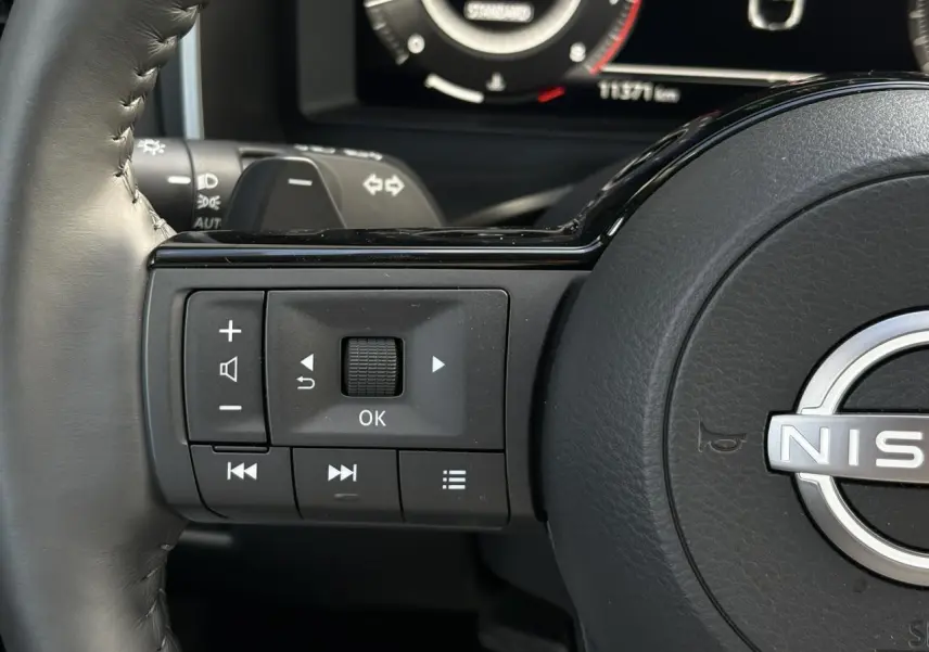 Gros plan sur les commandes gauche du volant cuir du Nissan Qashqai 2025, avec tableau de bord numérique en arrière-plan.