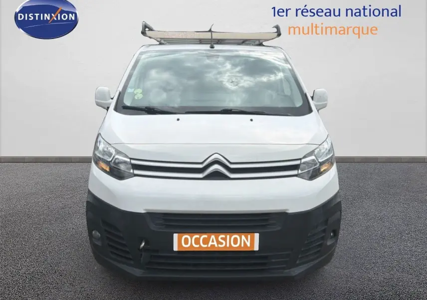 Vue avant d'un Citroën Jumpy XL blanc avec pare-chocs noir et galerie de toit, en arrière-plan un mur gris.