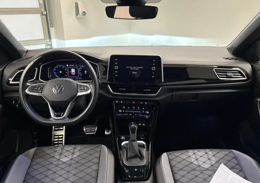 Vue intérieure avant du Volkswagen T-Roc 2024 avec tableau de bord moderne et sièges tissu gris à motifs.