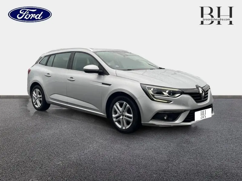 Renault Mégane Break gris platine vue 3/4 avant droit, avec phares LED et barres de toit visibles.