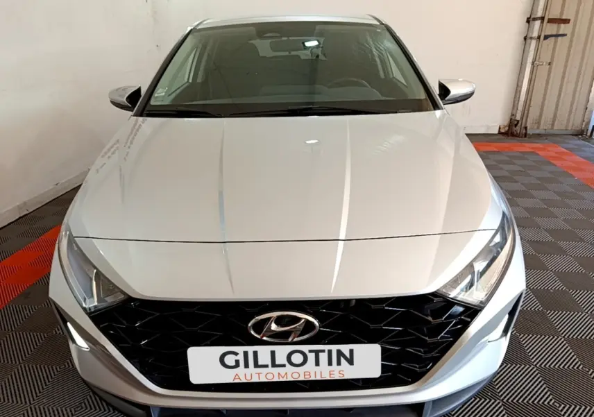Vue avant d'une Hyundai i20 gris clair 2022 avec calandre noire et phares LED, stationnée en intérieur.
