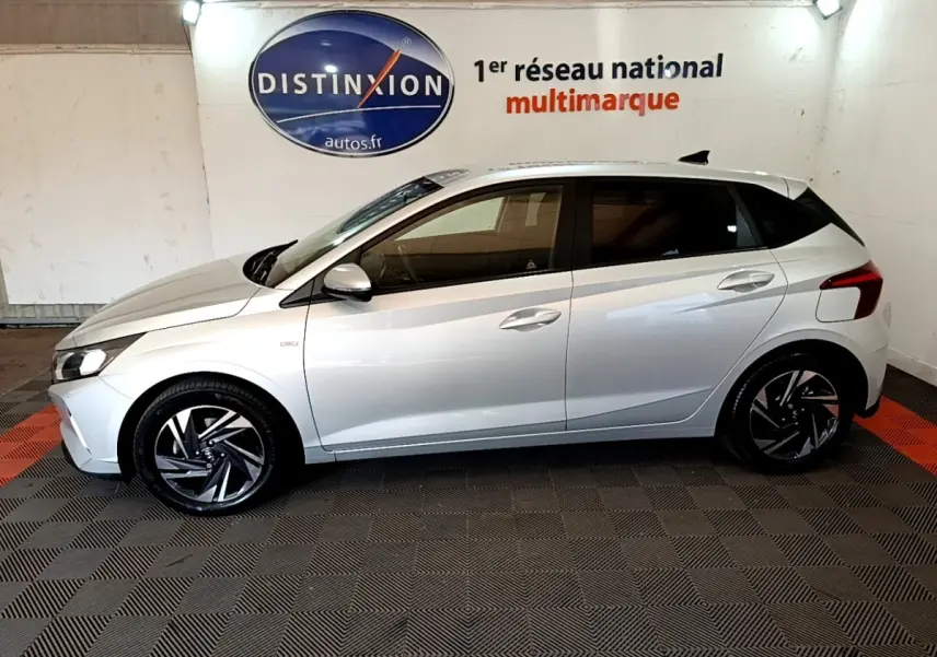 Vue de profil côté gauche d'une Hyundai i20 gris clair 2022 en intérieur avec jantes alliage noires et logo Distinxion en arrière-plan.