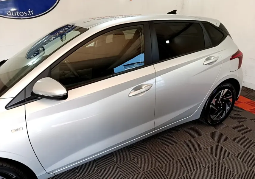 Vue côté gauche d'une Hyundai i20 gris clair 2022, mettant en valeur les jantes noires et les vitres teintées.