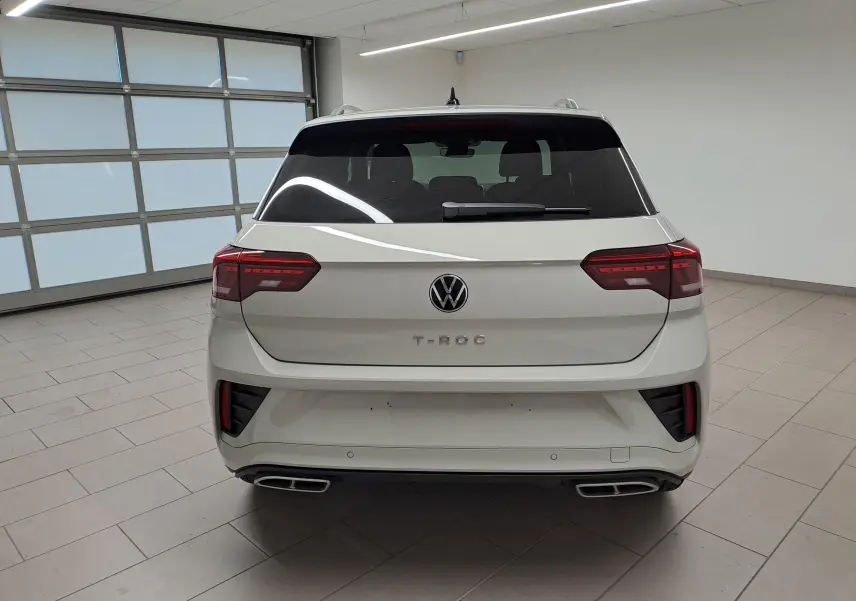 Vue arrière d'un Volkswagen T-Roc 2024 gris Ascot avec double sortie d'échappement et vitres teintées en intérieur showroom.