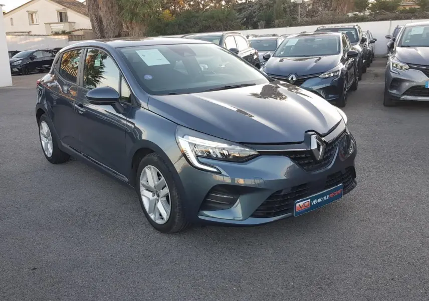 Renault Clio gris en vue 3/4 avant droit, avec phares allumés et jantes alliage visibles sur parking.