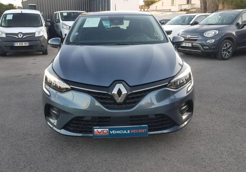 Vue frontale d'une Renault Clio gris métallisé avec feux allumés et plaque "Véhicule récent" sur un parking.