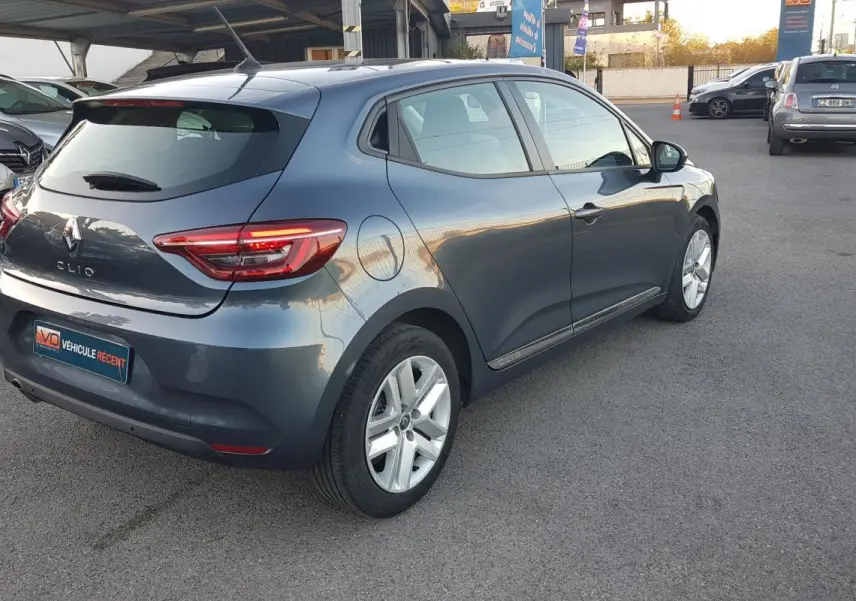 Renault Clio gris vue 3/4 arrière droit sur parking, avec feux arrière allumés et jantes alliage visibles.