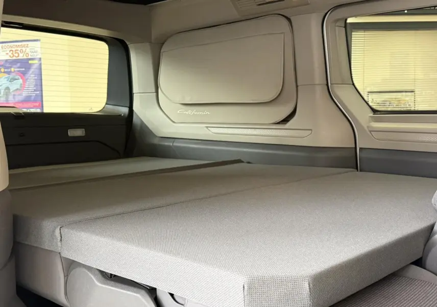 Vue intérieure arrière du Volkswagen California T7 blanc Candy montrant la couchette dépliée et la fenêtre latérale.