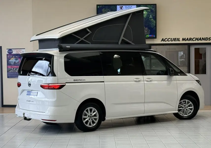 Vue latérale droite d’un Volkswagen California T7 blanc Candy avec toit relevable noir ouvert en intérieur showroom.