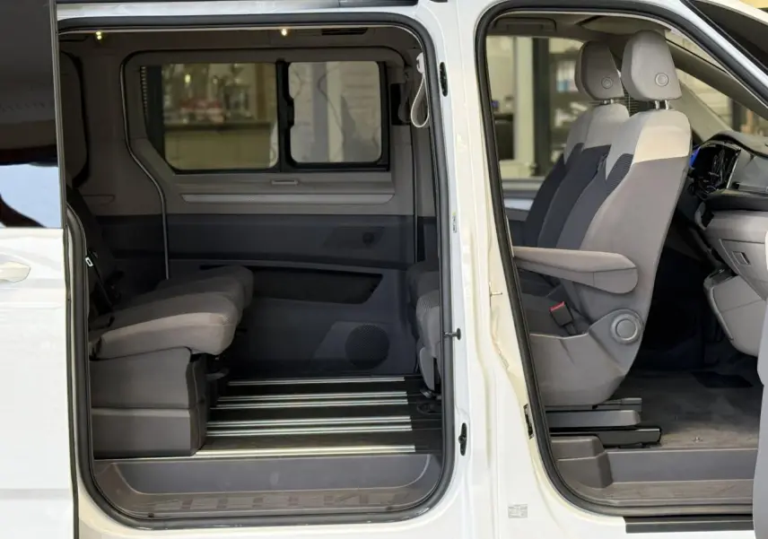 Vue latérale droite intérieure du Volkswagen California T7 blanc Candy montrant les sièges avant avec accoudoirs et la banquette arrière.