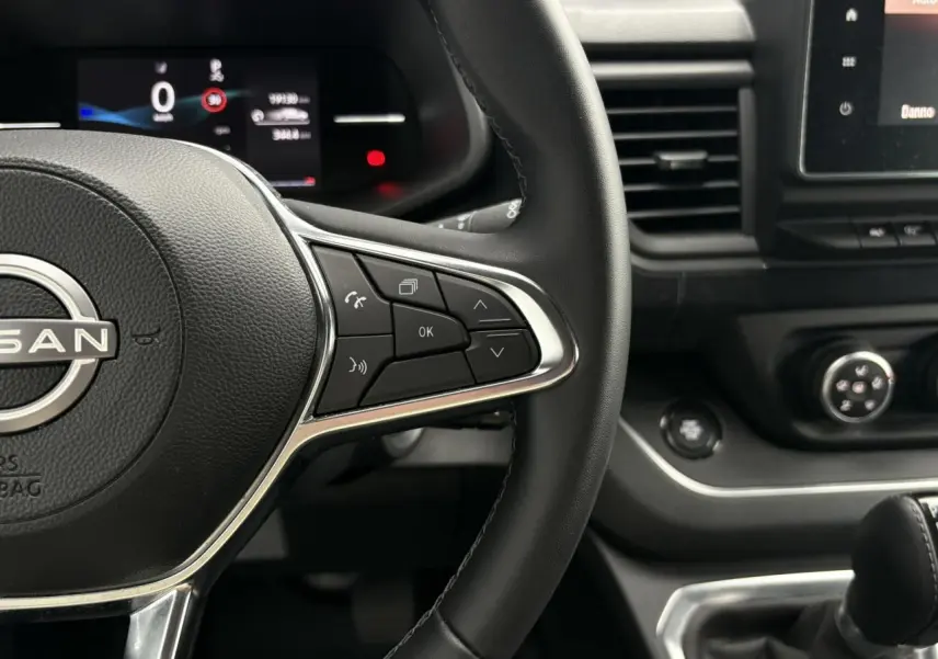Vue rapprochée du volant noir et du tableau de bord du Nissan Primastar gris Comète avec commandes au volant et écran multifonction.