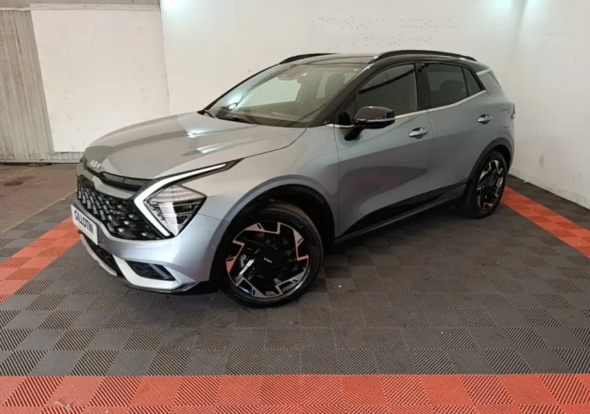 KIA Sportage gris clair en 3/4 avant droit, avec jantes noires et toit noir dans un garage éclairé.