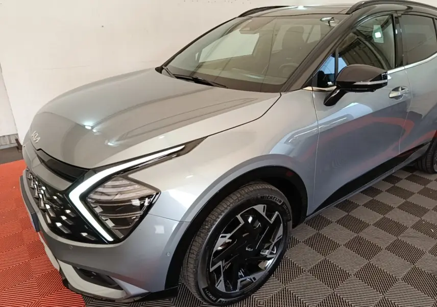 Vue 3/4 avant droit d’un KIA Sportage gris clair avec phares LED en forme de boomerang et jantes noires distinctives.
