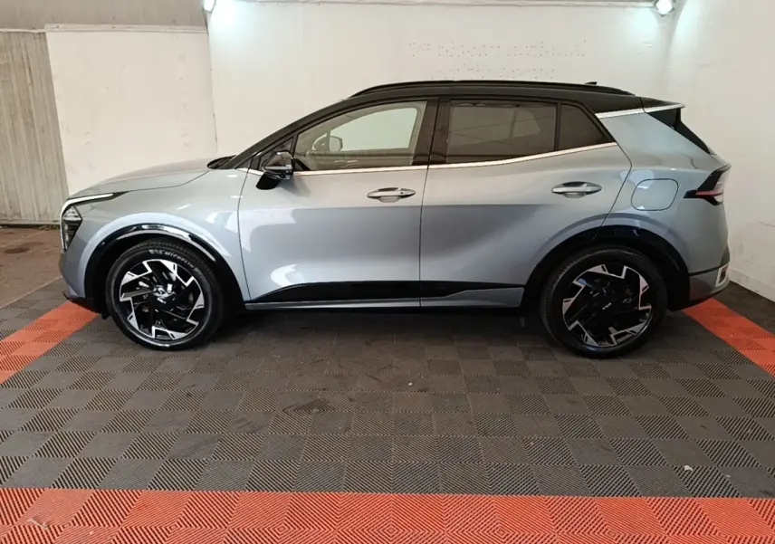 Vue de profil côté gauche d'un KIA Sportage gris clair avec toit noir et jantes noires stylisées, stationné en intérieur.