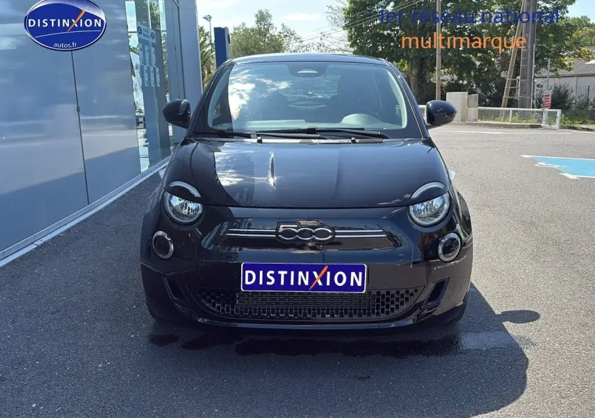 Vue avant d'une FIAT 500 électrique noire avec logo 500 chromé et phares ronds LED, stationnée devant un bâtiment.