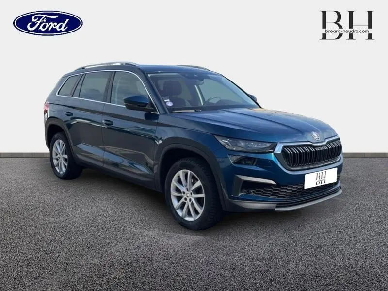 Vue 3/4 avant d'un Skoda Kodiaq bleu lave métallisé avec jantes alu et barres de toit visibles sur fond neutre.