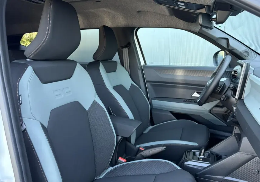 Vue intérieure côté conducteur du Dacia Duster 2026, sièges bi-ton gris clair et noir avec accoudoir central.