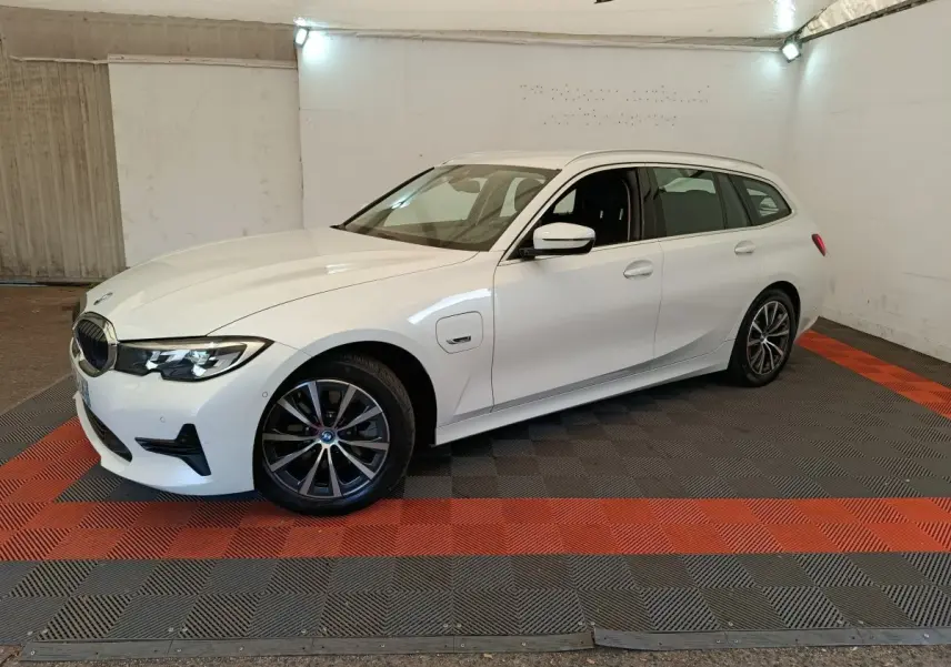 BMW Série 3 320e blanche vue 3/4 avant droit en break, avec jantes alliage et prise de recharge latérale visible.