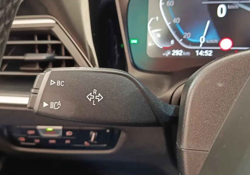 Gros plan sur la commande de clignotant gauche dans l’habitacle d’une BMW Série 3 hybride blanche, tableau de bord numérique visible.
