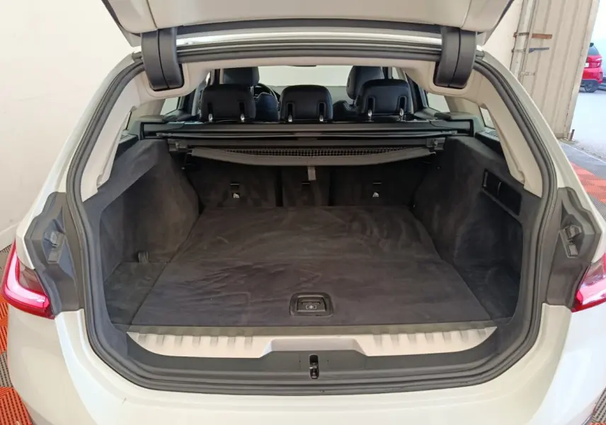 Coffre ouvert vue arrière d’une BMW Série 3 320e blanche, espace de chargement avec tapis noir et banquette rabattable.