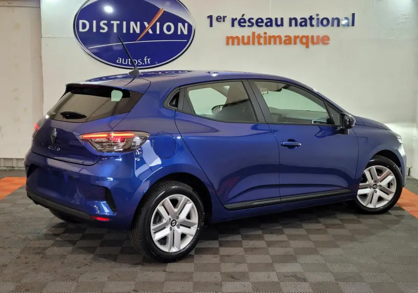 Vue 3/4 arrière droite d'une Renault Clio bleu 2025 stationnée en intérieur avec feux arrière allumés.