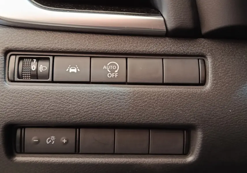 Gros plan sur les boutons de commande du tableau de bord noir du Nissan Qashqai 1.3 MHEV gris foncé, avec régulateur de luminosité et aides à la conduite.