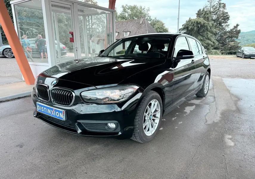 BMW Série 1 noire vue en 3/4 avant droit, avec calandre caractéristique et jantes argentées sous un abri.