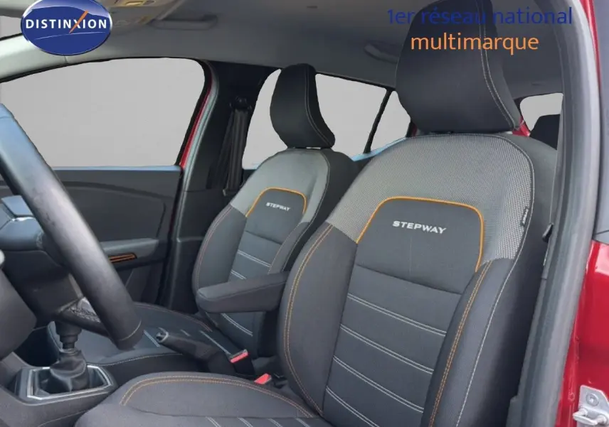 Vue intérieure côté conducteur de la Dacia Sandero Stepway rouge fusion, mettant en valeur les sièges noirs avec surpiqûres orange et logo Stepway.