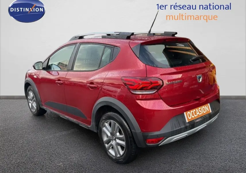 Vue 3/4 arrière droite d'une Dacia Sandero Stepway rouge fusion avec barres de toit et protections noires sur les bas de caisse.